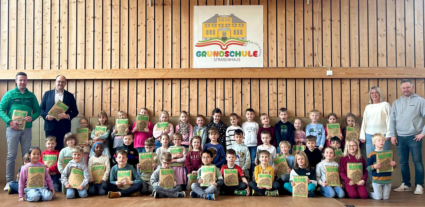 A large group of children and five adults pose indoors, holding what appear to be books or certificates, in front of a wooden wall with a sign that reads Grundschule Strassenhaus and a house illustration.